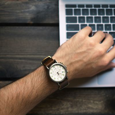 The left hand of a white man wearing a watch on the keyboard of a laptop.  The right hand is slightly in view. 